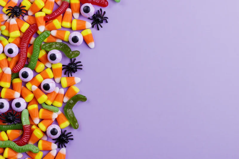 Assorted halloween candy scattered along left edge of purple surface with copyspace