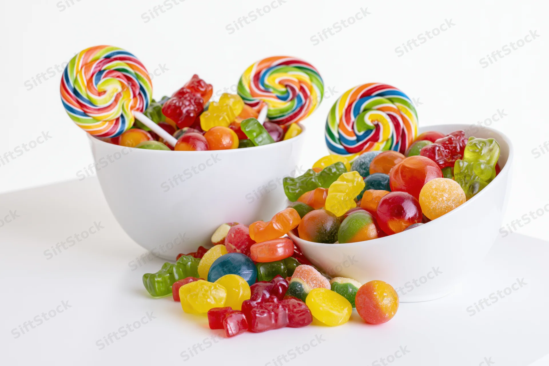 Two white ceramic bowls filled with colorful assorted candies including lollipops and gummy bears, some spilled onto the surface