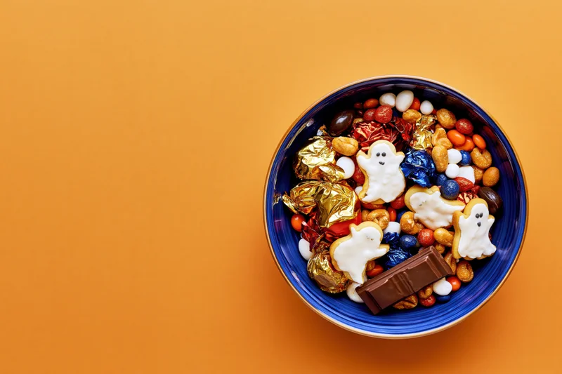 Blue ceramic bowl filled with halloween candy and ghost-shaped cookies on orange background