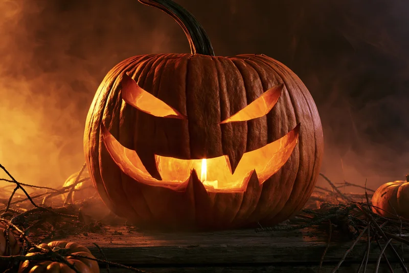 Carved pumpkin glowing from within on a dark smoky background with branches and smaller pumpkins around
