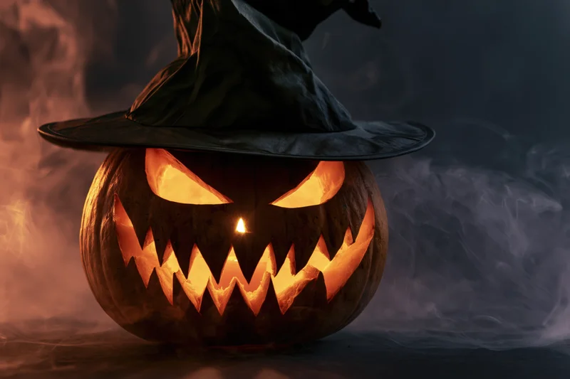 Carved jack-o-lantern with glowing eyes and jagged grin wearing a black witch hat in smoky dark setting