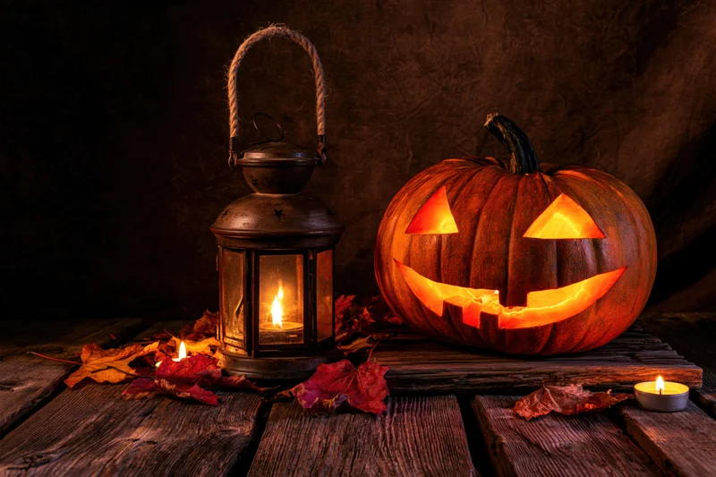 Carved jack-o-lantern with glowing eyes and a lit lantern on a wooden table surrounded by autumn leaves
