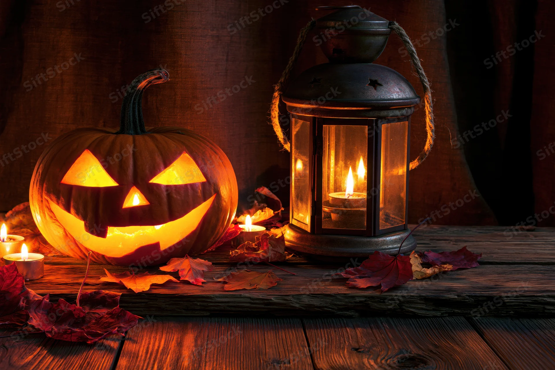 Carved jack-o-lantern glowing beside a lit lantern on a wooden table with autumn leaves