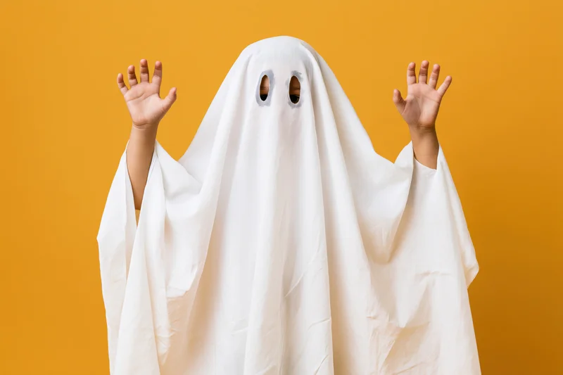 Child in white sheet ghost costume with hands raised against orange background
