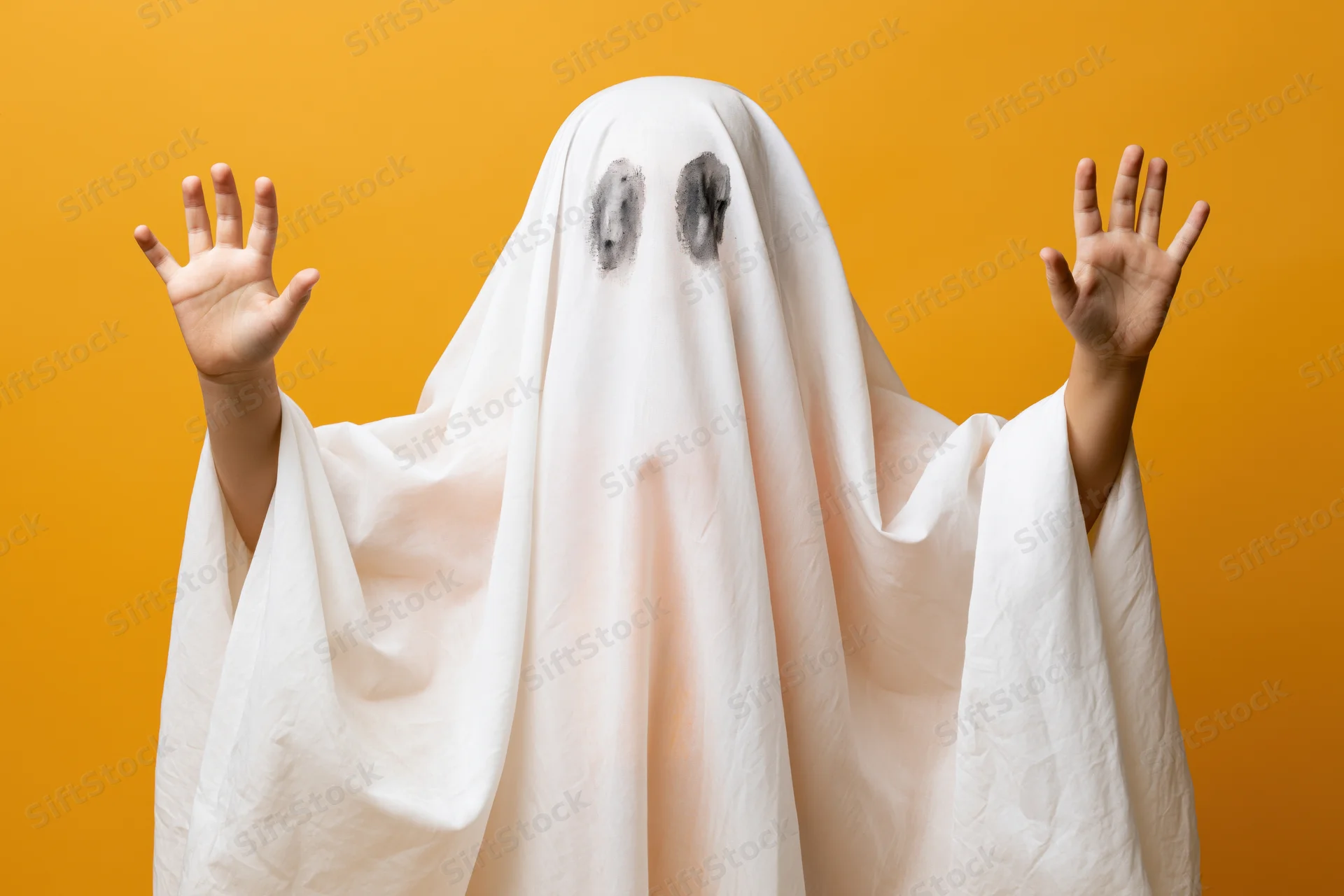 Child in white sheet ghost costume with hands raised against orange background
