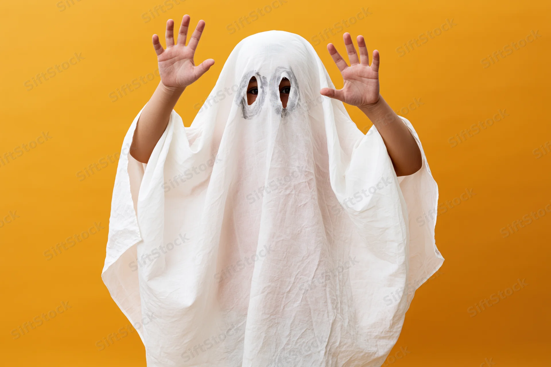 Child in white sheet ghost costume with hands raised against orange background