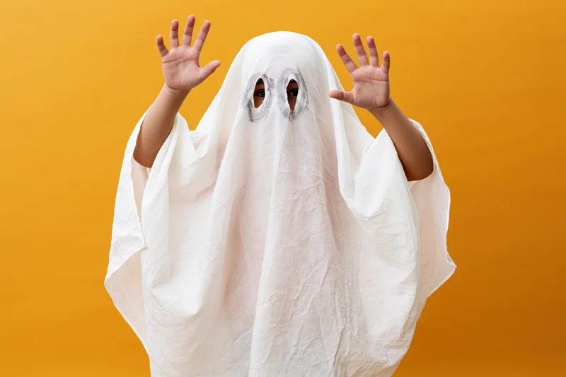 Child in white sheet ghost costume with hands raised against orange background