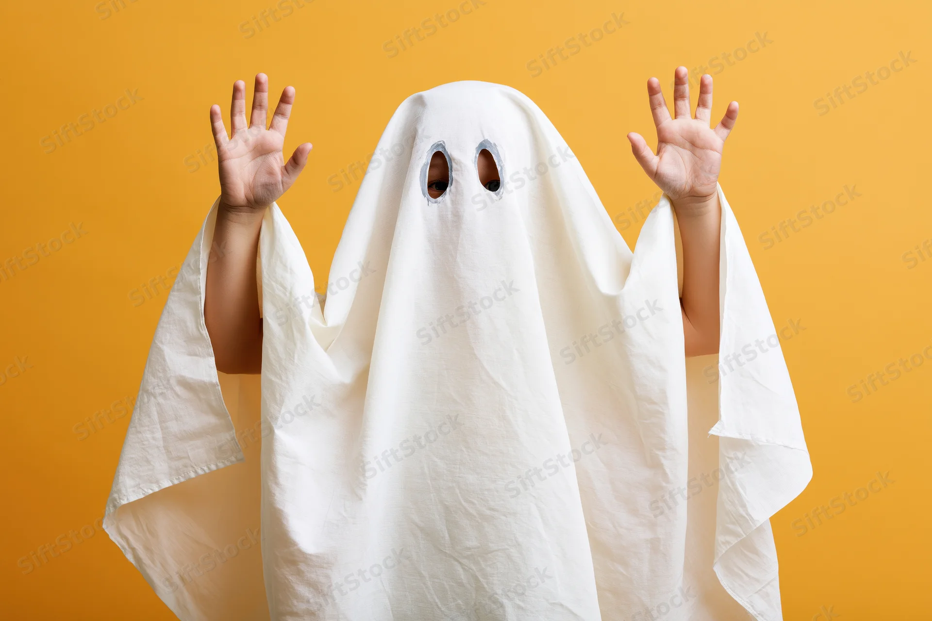 Child in white sheet ghost costume with hands raised against orange background