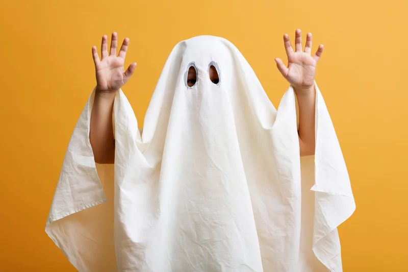Child in white sheet ghost costume with hands raised against orange background