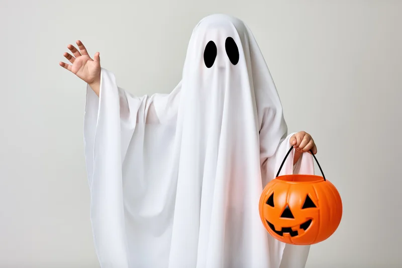 Child in white sheet ghost costume waving and holding orange pumpkin bucket for trick or treat