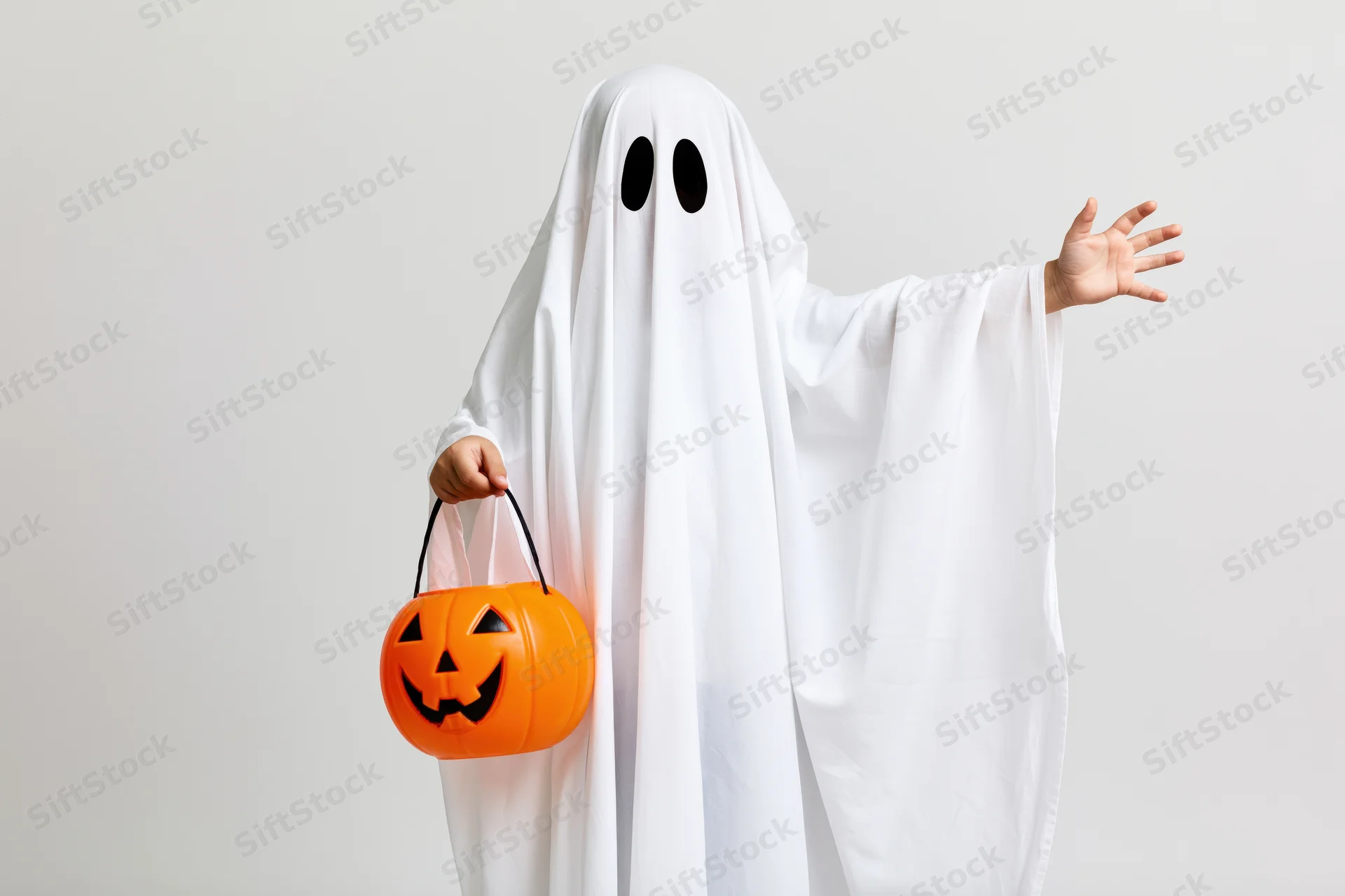 Child in ghost costume holding orange pumpkin bucket and reaching out against plain background