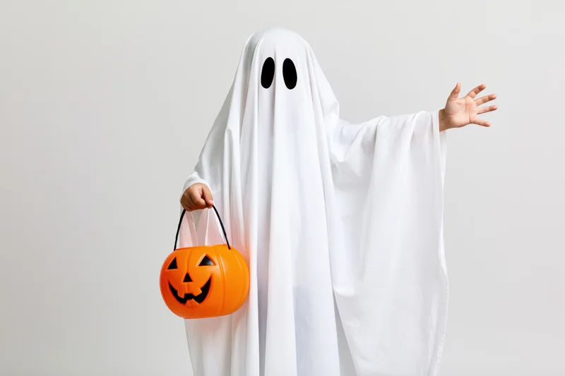 Child in ghost costume holding orange pumpkin bucket and reaching out against plain background