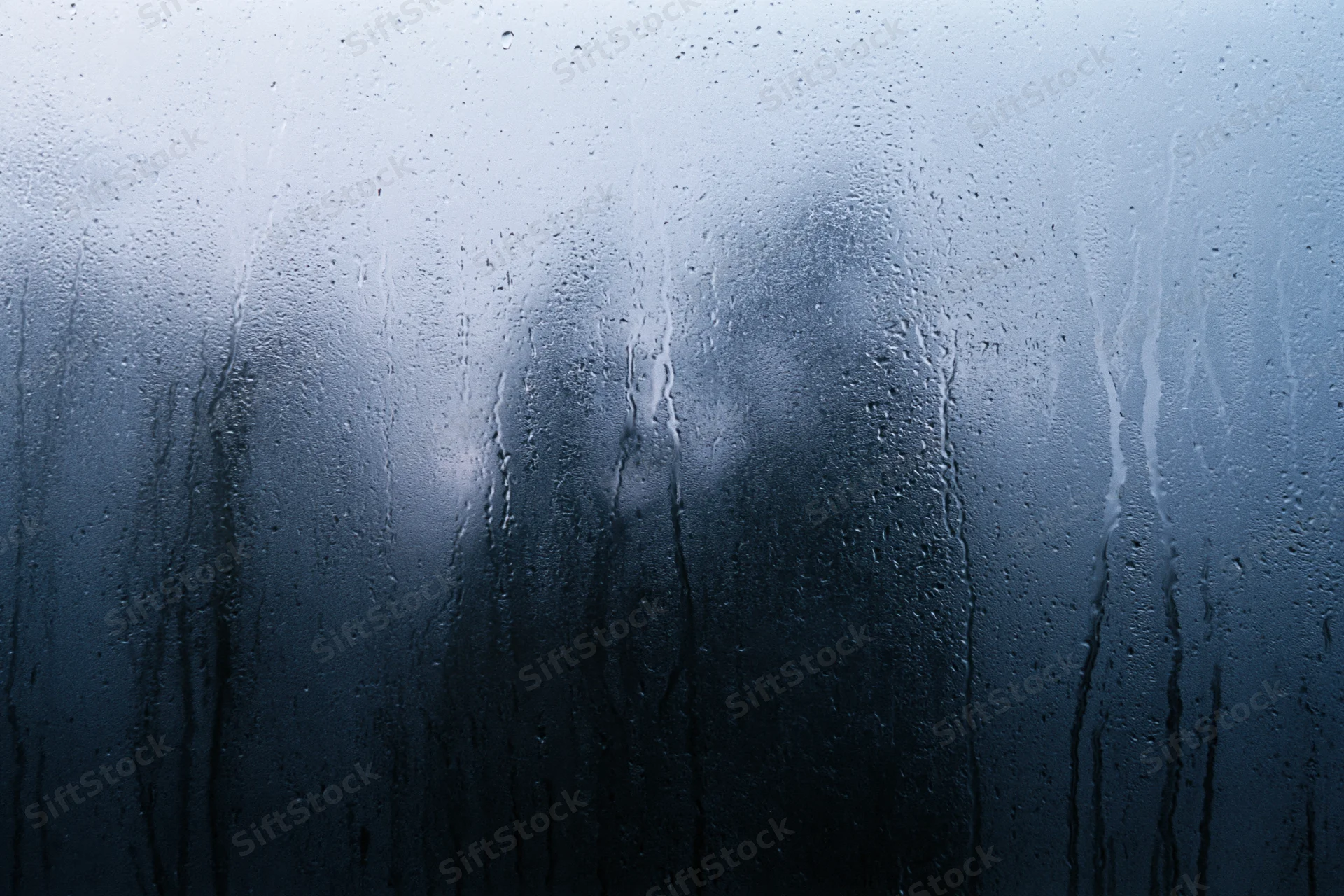 Rain-streaked window glass with water droplets and fog, showing blurred reflection in dark blue tones