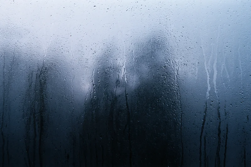 Rain-streaked window glass with water droplets and fog, showing blurred reflection in dark blue tones