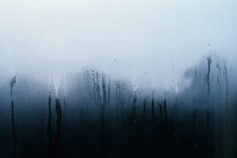 Rain-streaked window glass with condensation and water droplets, showing blurred outdoor view through fogged surface