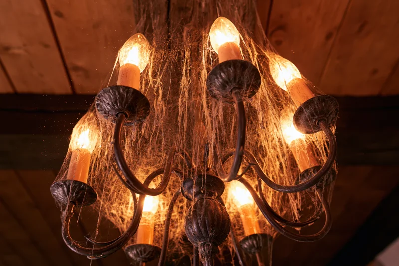 Old metal chandelier covered in cobwebs glowing warmly against wooden ceiling