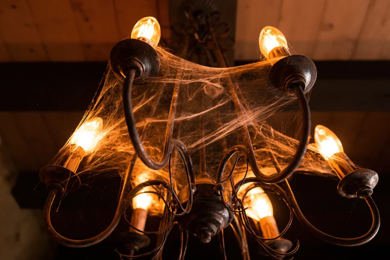 Old metal chandelier with glowing bulbs and spiderwebs hanging from ceiling indoors