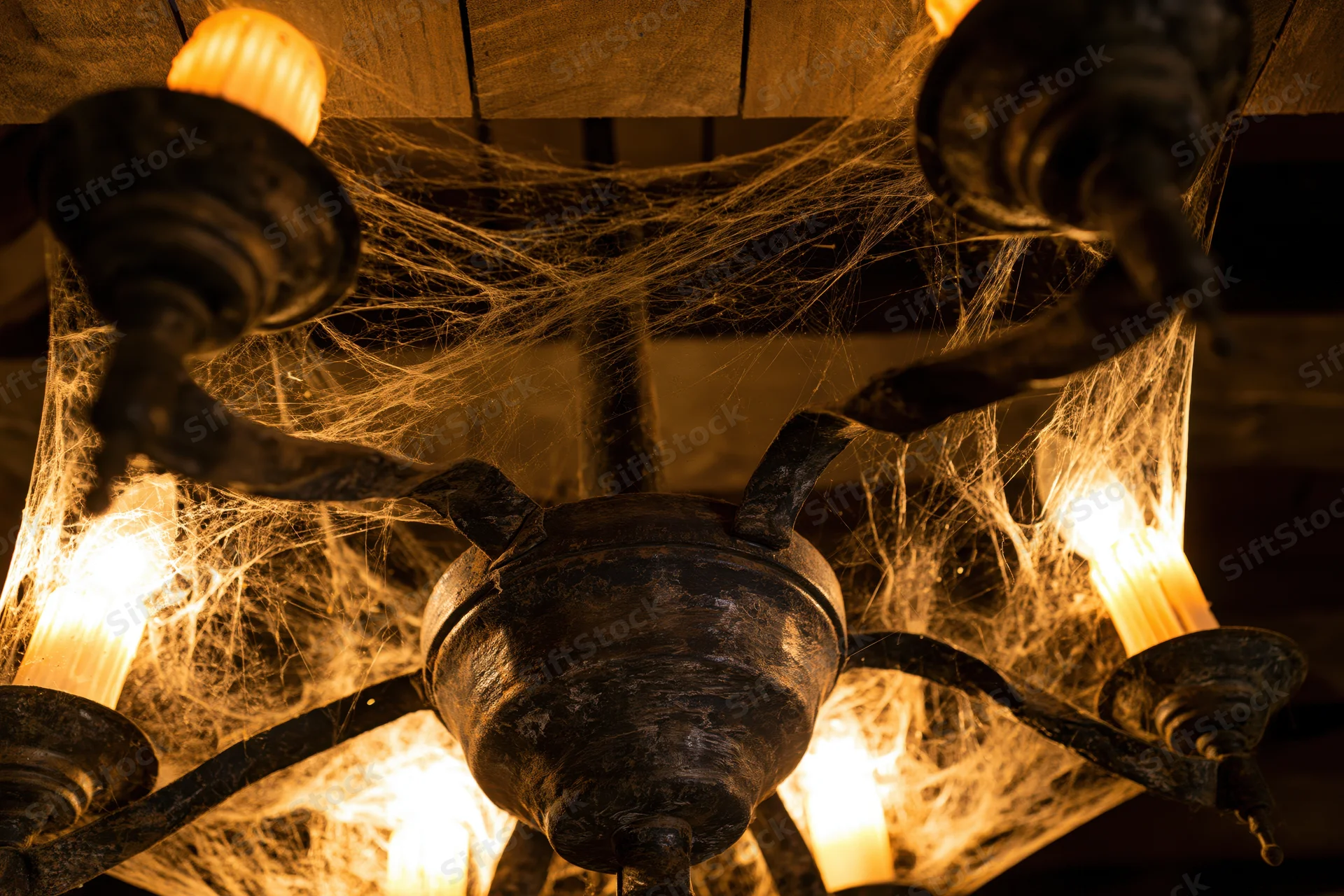 Old metal chandelier covered in cobwebs with glowing bulbs hanging from wooden ceiling