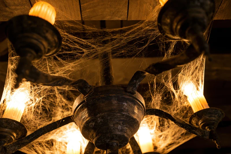 Old metal chandelier covered in cobwebs with glowing bulbs hanging from wooden ceiling