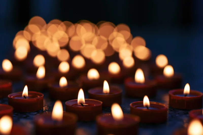 Dozens of small red tea light candles burning steadily arranged in rows on a dark surface with blurred lights in the background