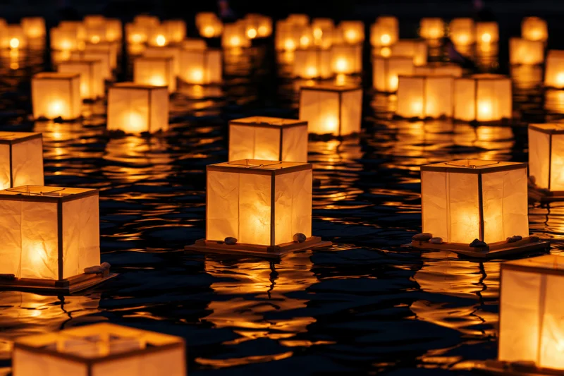 Square paper lanterns floating on dark water at night, glowing warmly with reflections rippling on the surface