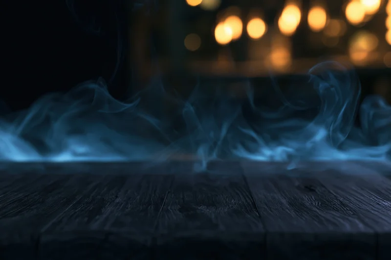 Dark wooden table surface with blue smoke rising in dimly lit interior, warm bokeh lights in background