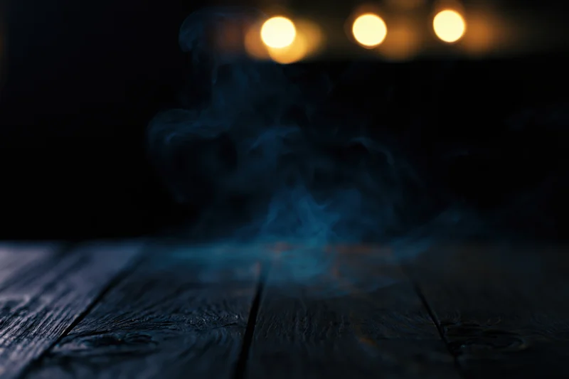 Smoke rising from a dark weathered wooden table surface at night with warm bokeh lights in the background