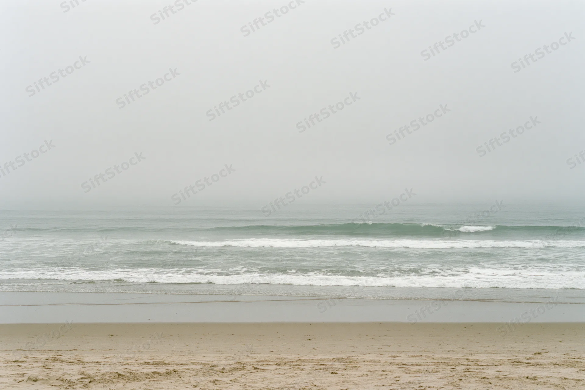 Foggy ocean shoreline with small waves breaking gently onto sandy beach under overcast sky