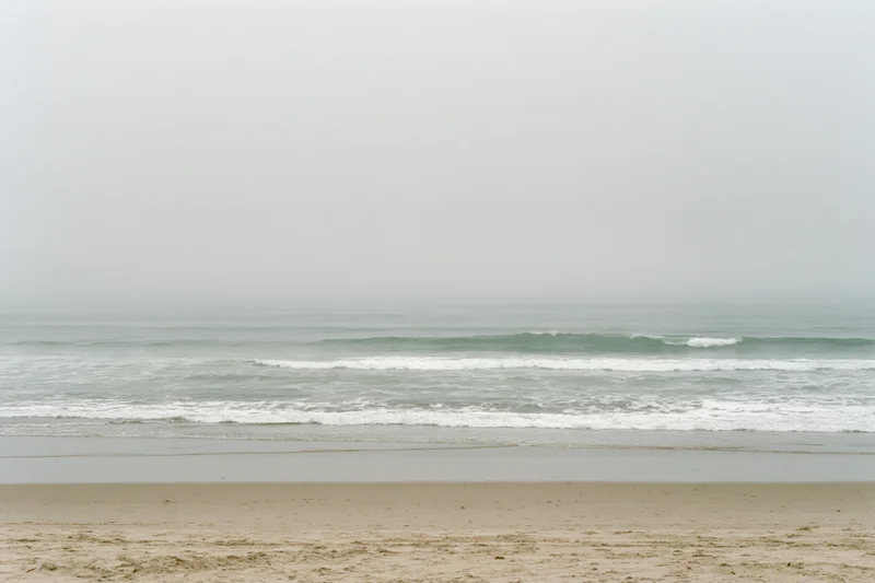 Foggy ocean shoreline with small waves breaking gently onto sandy beach under overcast sky