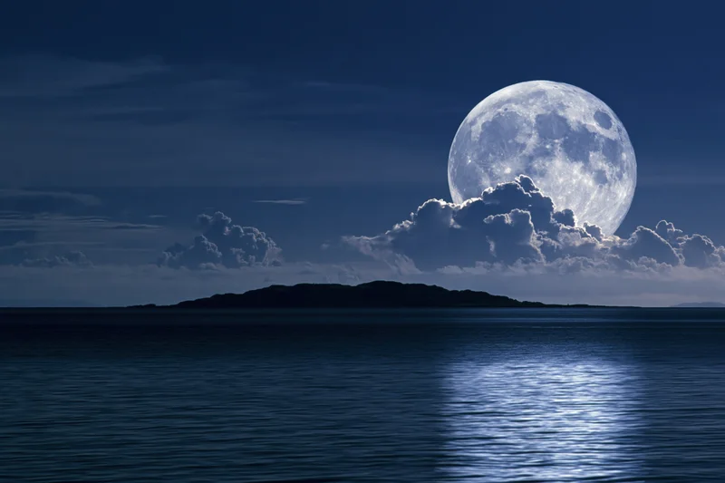 Full moon rising over calm ocean at night with clouds and reflection on water