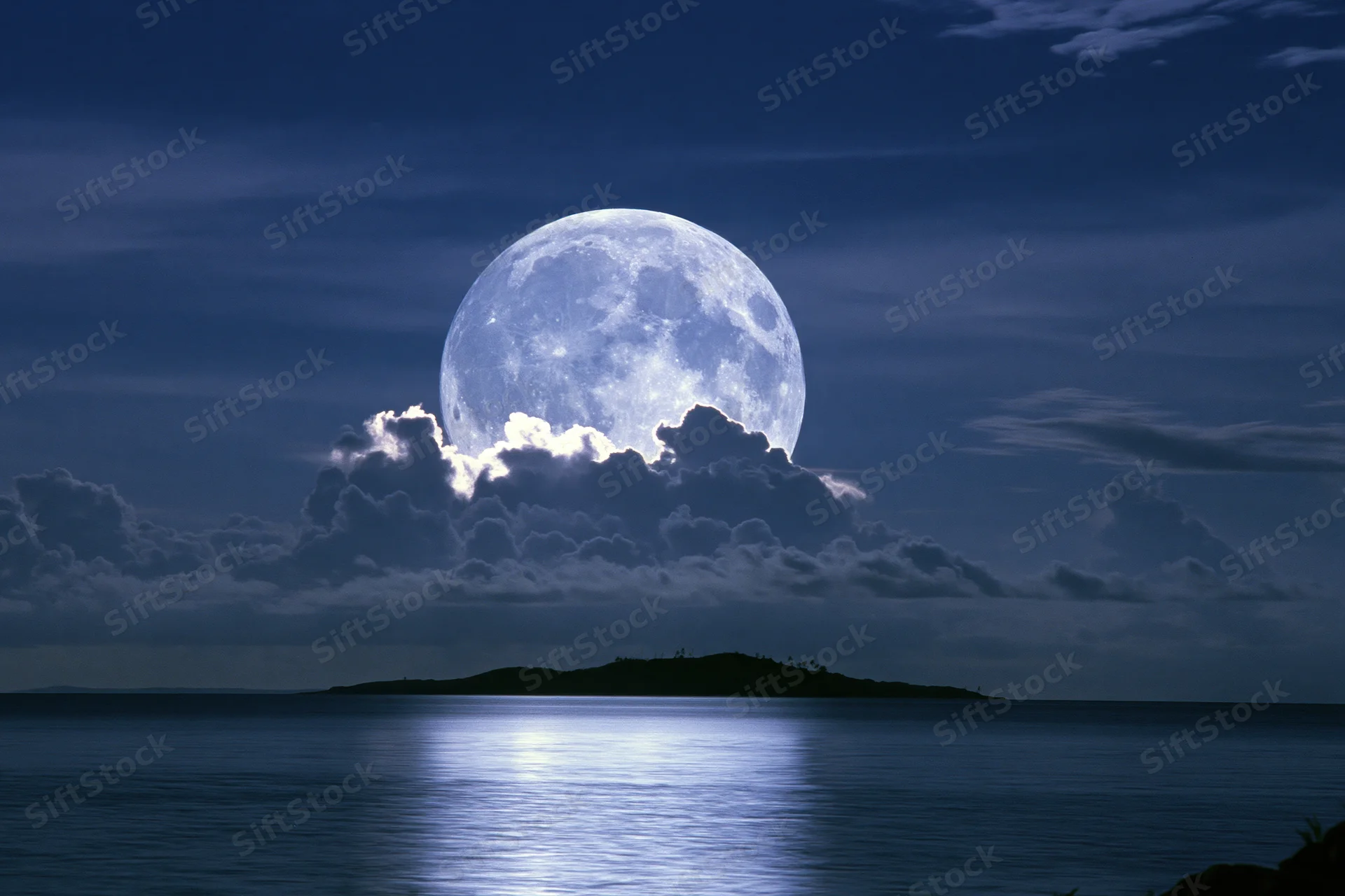 Full moon rising over calm ocean at night with clouds and island silhouette