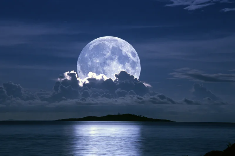 Full moon rising over calm ocean at night with clouds and island silhouette