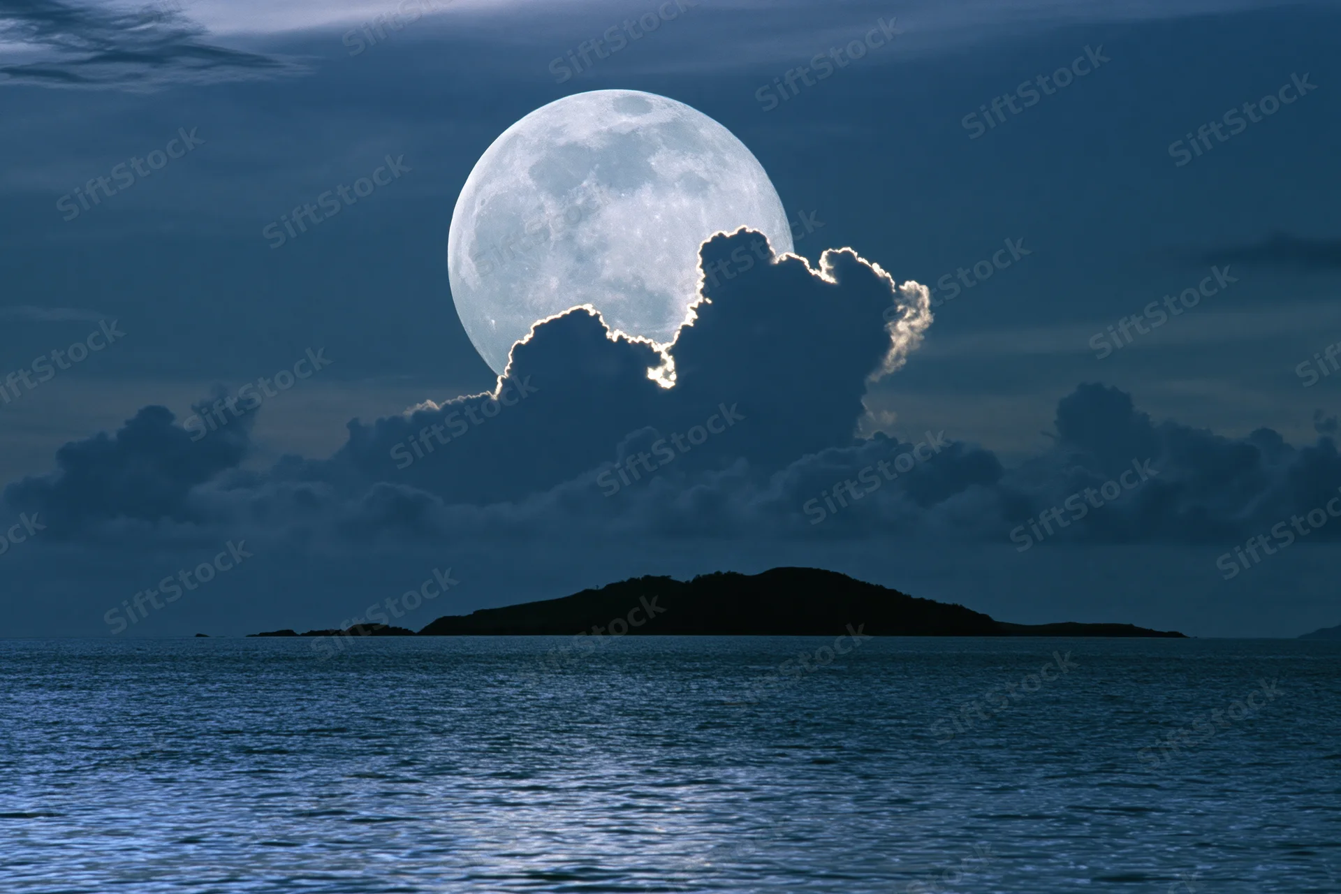 Full moon rising over calm ocean at night with clouds and island silhouette