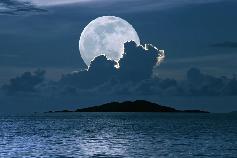 Full moon rising over calm ocean at night with clouds and island silhouette