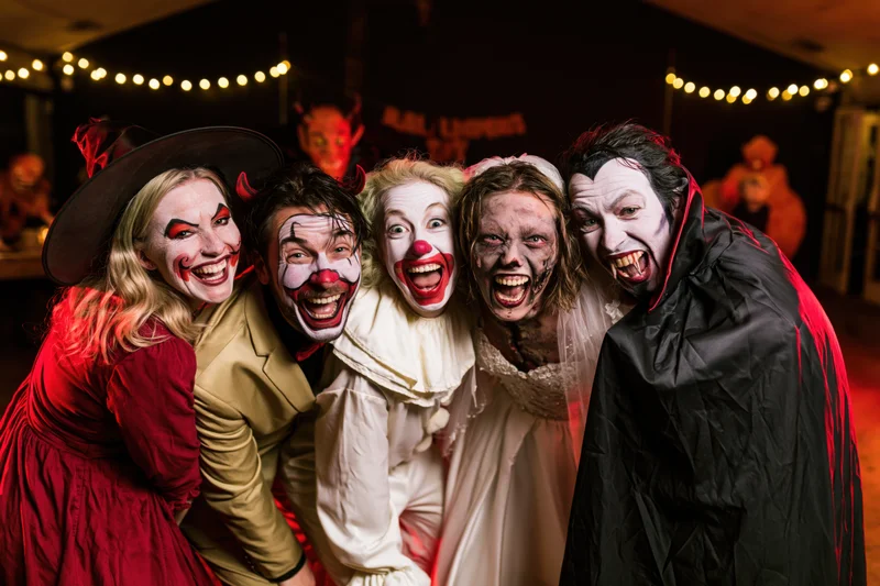 Five adults in halloween costumes posing together indoors with string lights and decorations