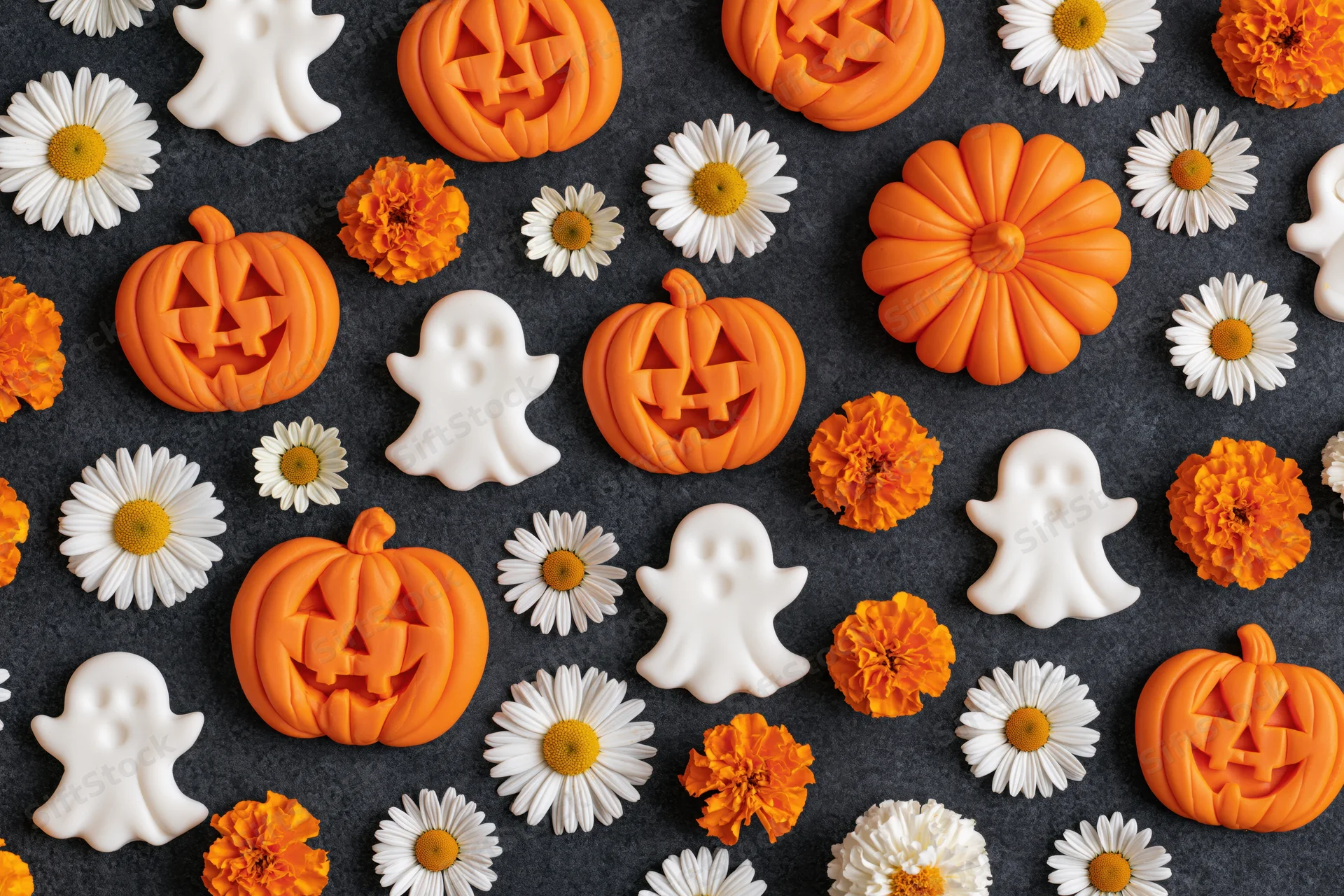 Carved jack-o-lanterns and white ghost figurines arranged with daisies and marigolds on dark surface