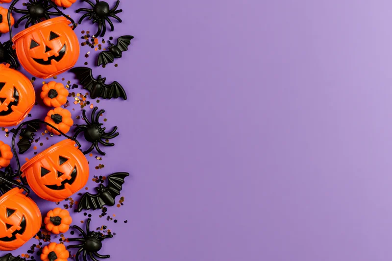 Orange plastic jack-o-lantern candy buckets and black spiders bats on purple surface with copyspace
