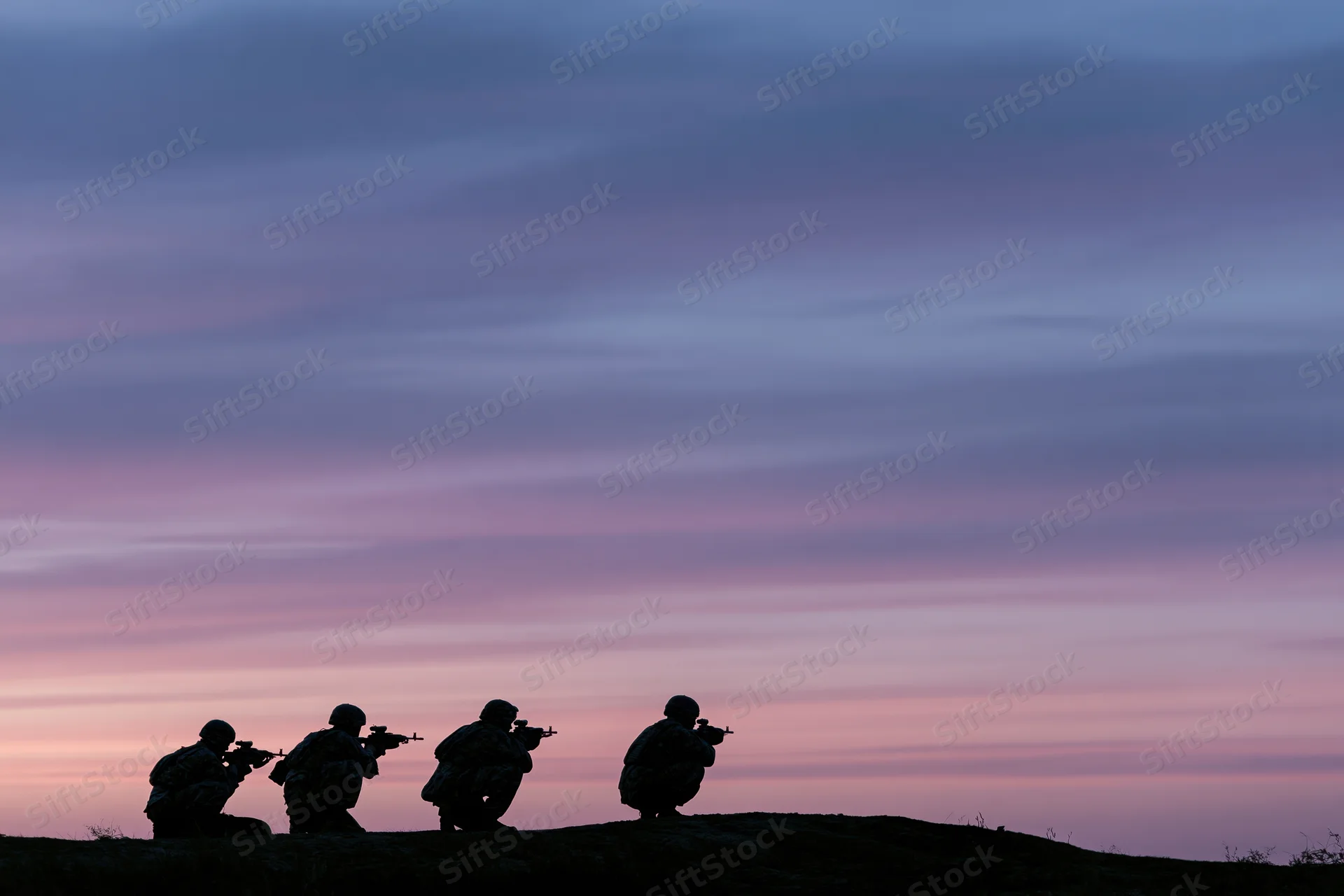 Four soldiers in combat gear crouching on a hill at twilight against a pink and purple sky