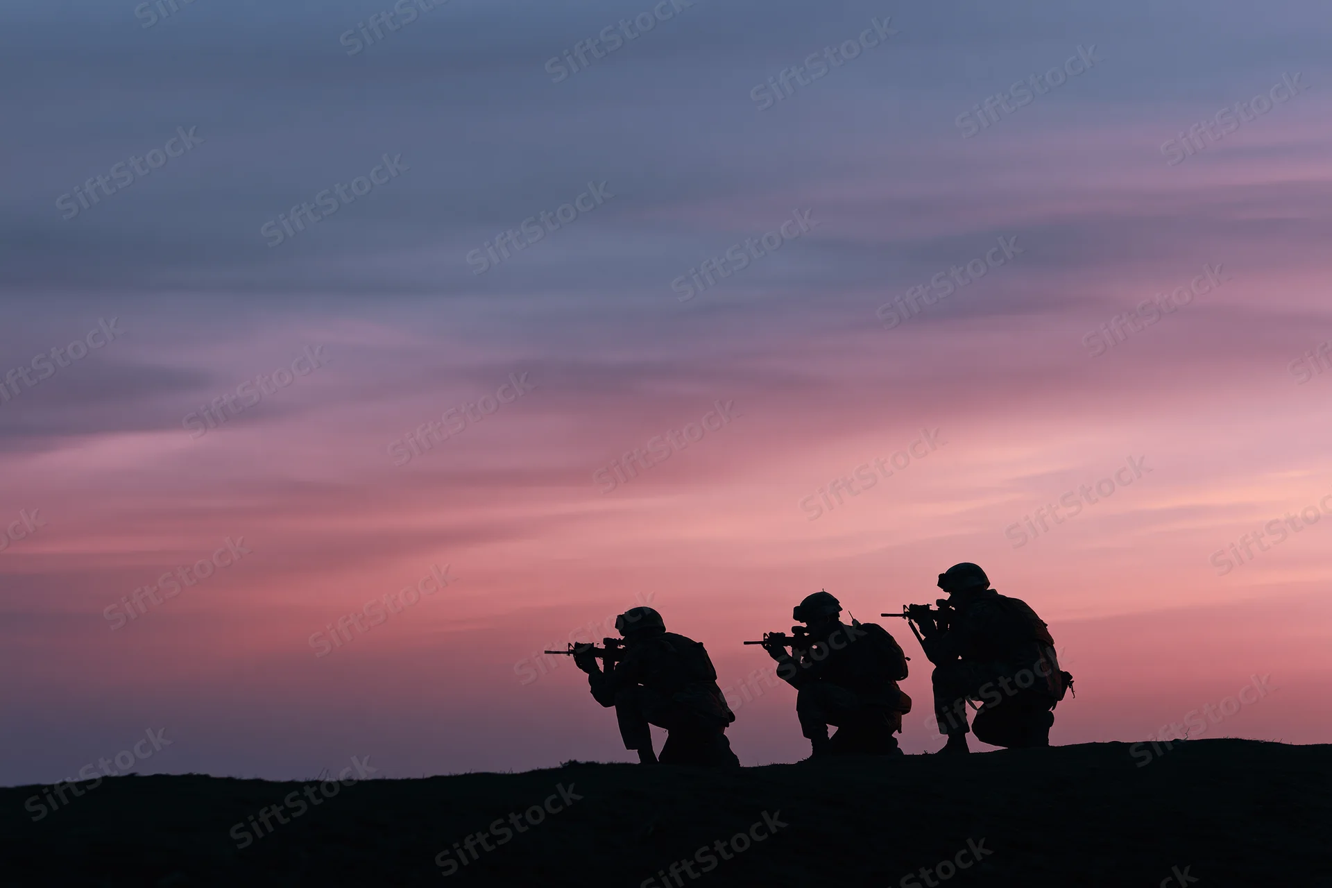 Three soldiers in combat gear crouching with rifles on a hill at dusk