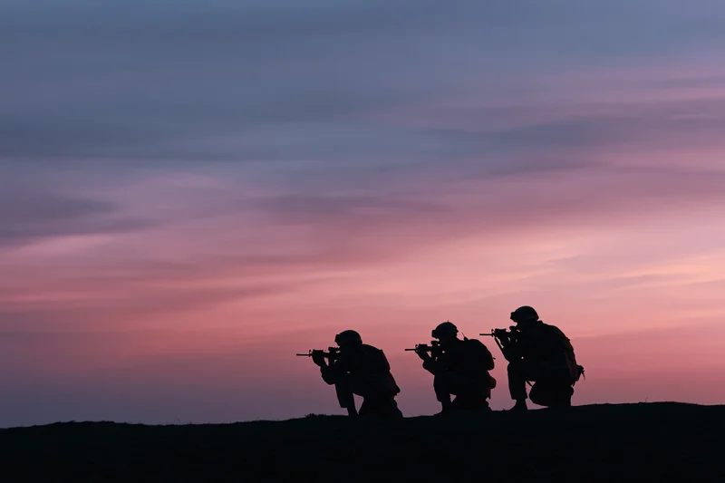 Three soldiers in combat gear crouching with rifles on a hill at dusk