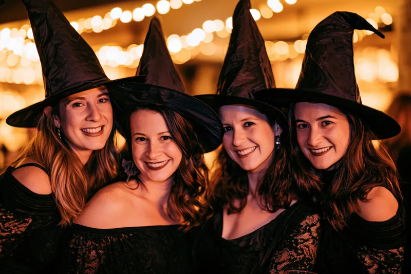 Four young women in black witch costumes smiling together at a halloween party