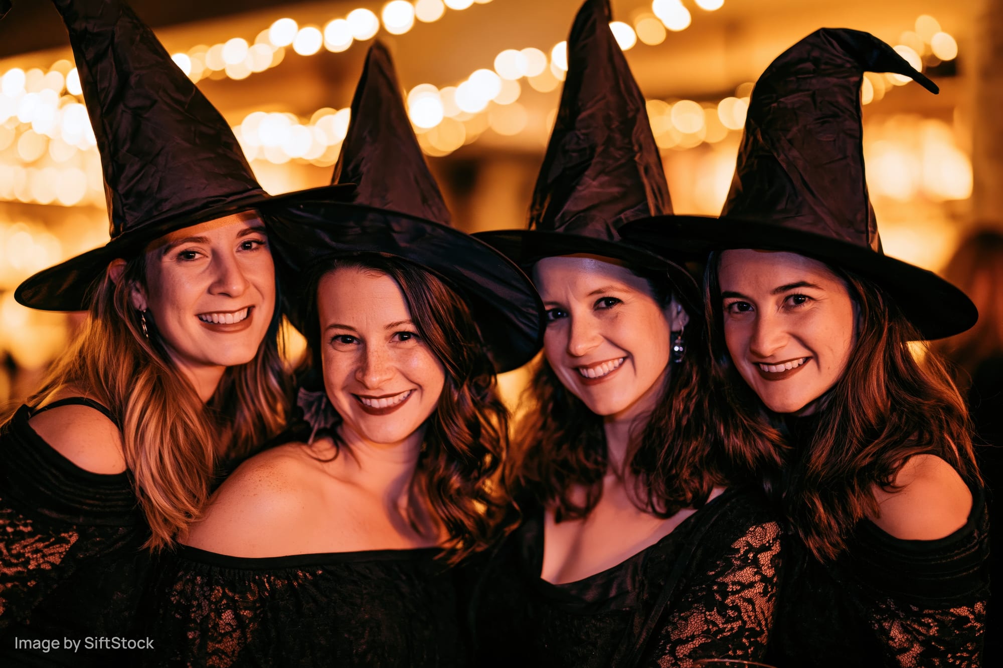Four young women in black witch costumes smiling together at a halloween party