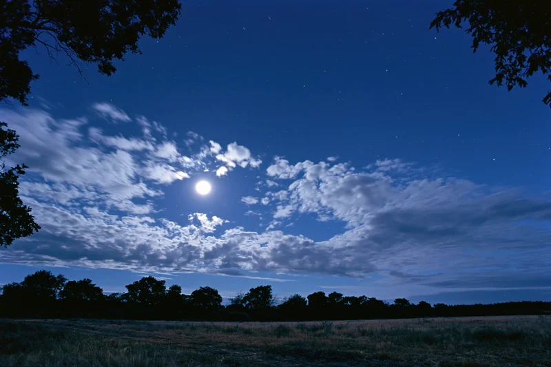 Full moon glowing among scattered stars and drifting clouds over a dark rural field with tree silhouettes