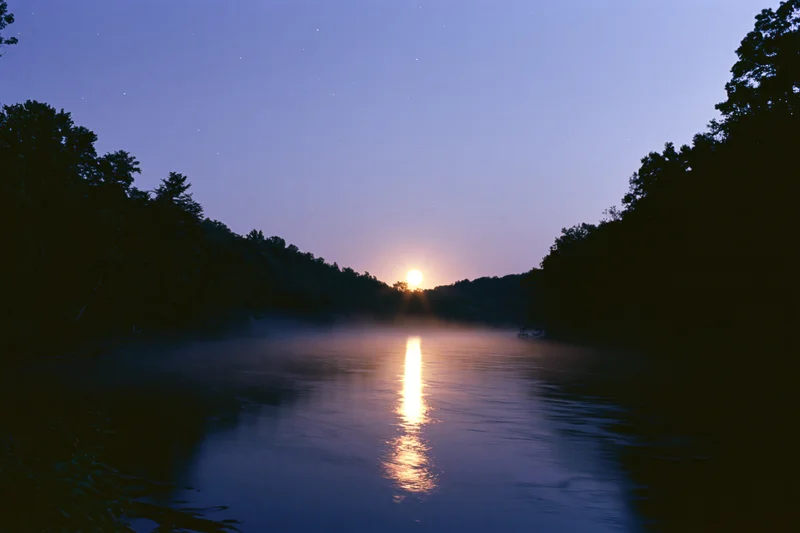 Full moon rising over calm river at night with reflection on water between tree-lined banks