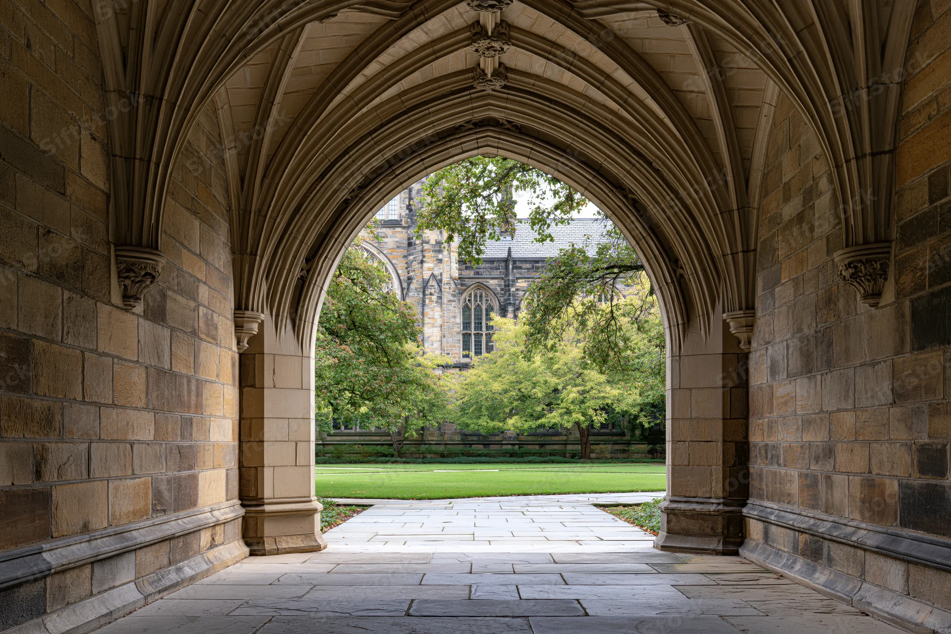 Gothic stone archway with ribbed vaulted ceiling framing a view of a green courtyard and historic building beyond
