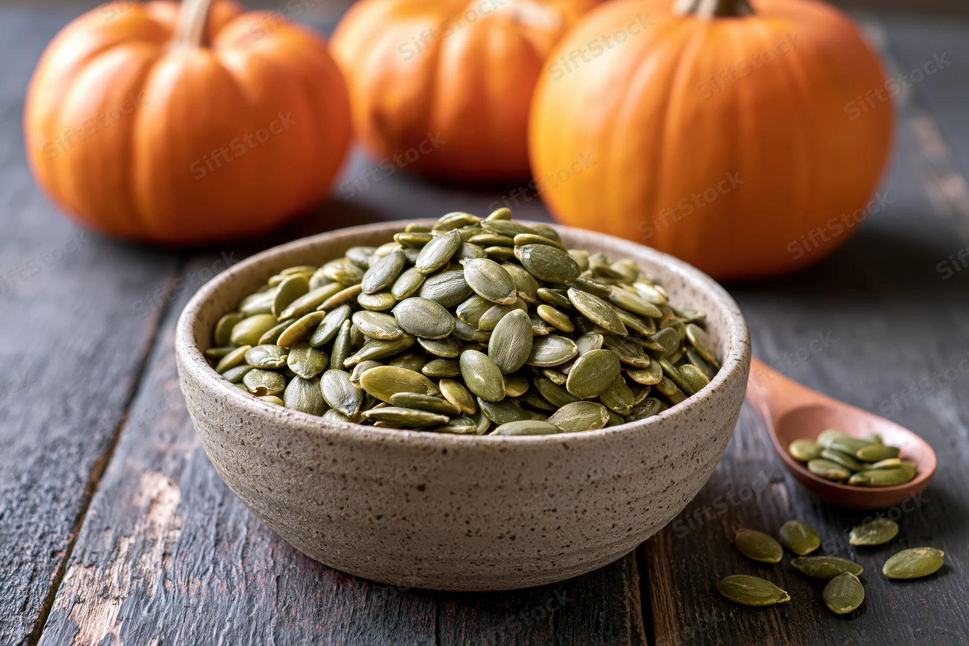 Green pumpkin seeds in a ceramic bowl on dark wood table with pumpkins in background
