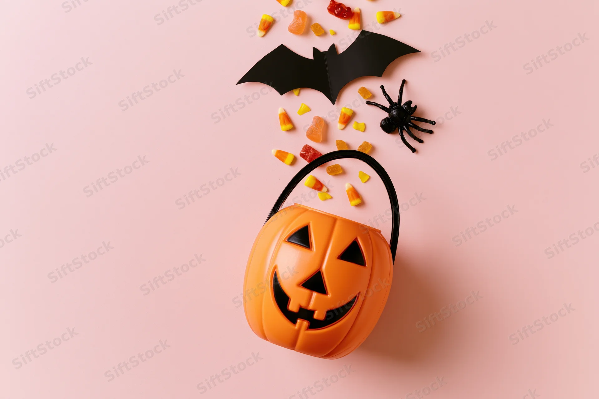 Jack-o-lantern candy bucket with scattered candy, bat cutout and spider on pink background