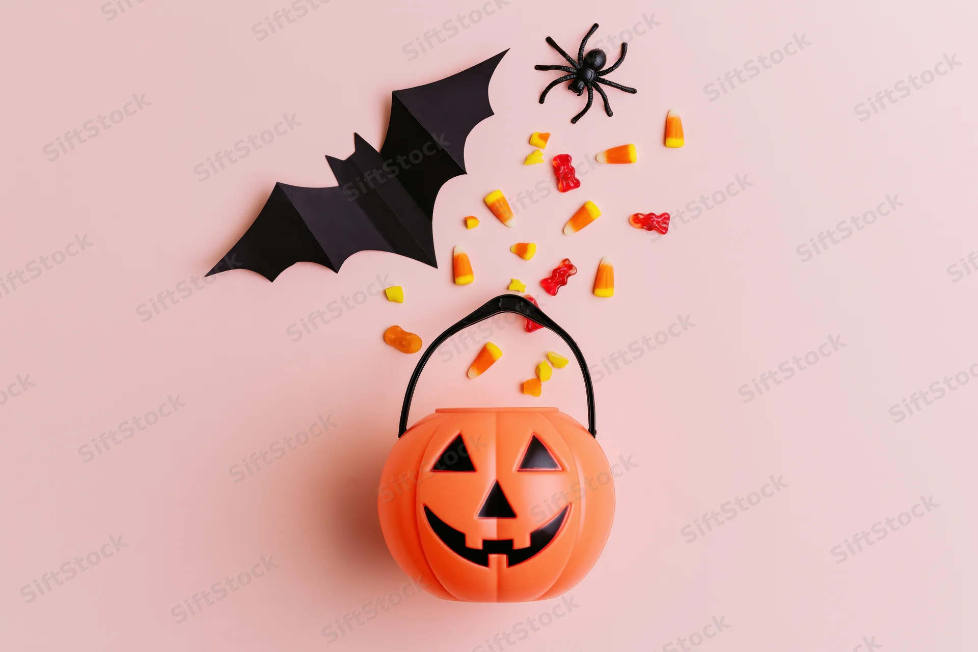Orange jack-o-lantern plastic candy bucket with scattered candy corn, gummy bears, paper bat, and toy spider on pink background