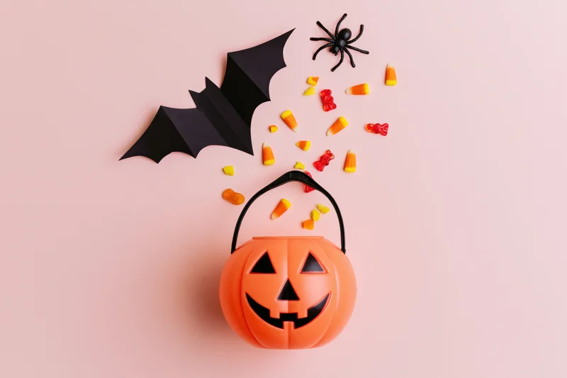 Orange jack-o-lantern plastic candy bucket with scattered candy corn, gummy bears, paper bat, and toy spider on pink background