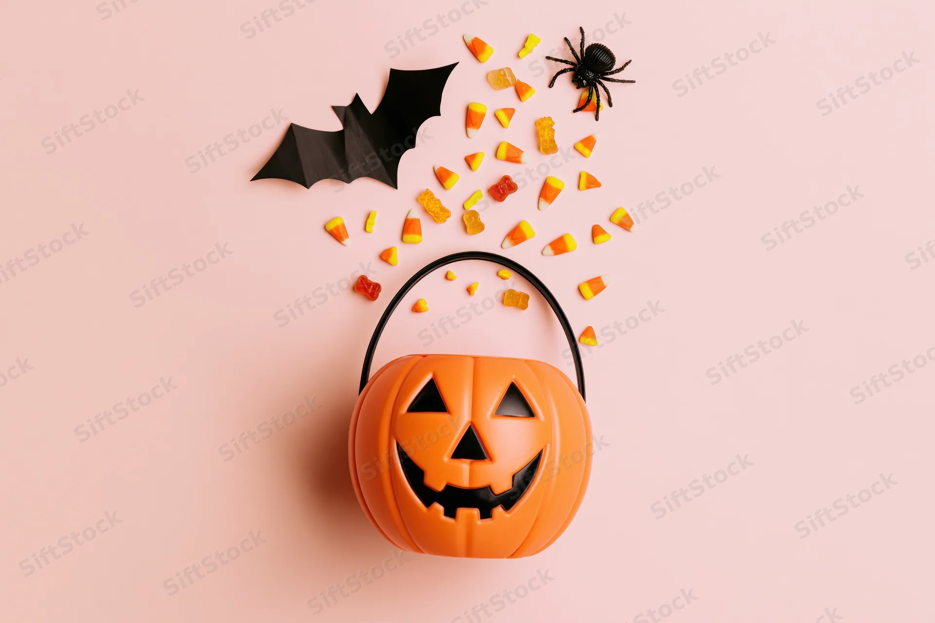 Jack-o-lantern candy bucket with scattered candy, bat, and spider on a pink background